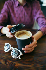 Female hands holding a cup of cappuccino with thick foam in black takeaway paper cup. Coffee take away.