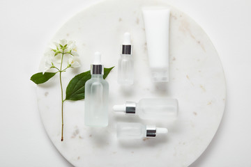 top view of cream tube and cosmetic glass bottles in plate with jasmine on white surface - obrazy, fototapety, plakaty
