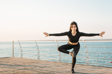 Pretty young brunette woman in a black jumpsuit practicing yoga on a pier at sunrise.