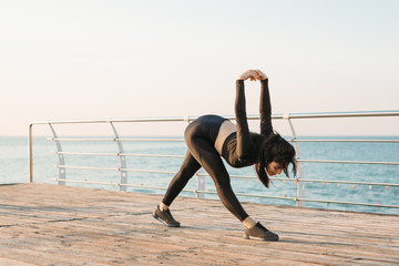 Young brunette woman with white wireless headphones in a black jumpsuit takes exercises on a pier at sunrise.