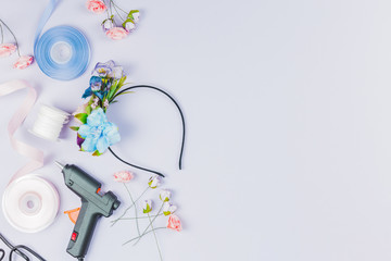 An overhead view of blue and white ribbon; artificial flower; glue gun for making hairband on white backdrop