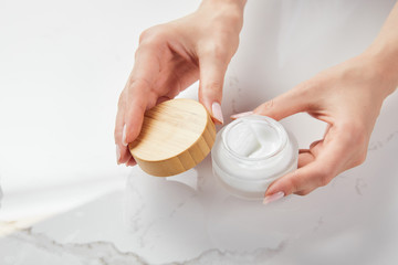 cropped view of woman opening jar with cream on white surface