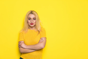 Portrait of a smiling blonde young woman standing with arms folded isolated over yellow background