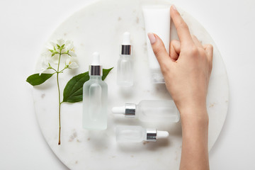 Cropped view of woman hand with cream tube over plate with jasmine and cosmetic glass bottles on white surface - obrazy, fototapety, plakaty