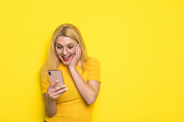 Fototapeta premium Portrait of shocked young blonde girl looking at mobile phone isolated over yellow background