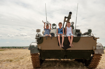 Fototapeta premium Three children in colorful clothes climbed onto modern battle tank. Children play while no tank crews. concept of contrasting innocent childhood and war