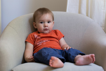 Horizontal full length portrait of cute fair toddler girl in orange shirt and blue pants lounging in armchair with adorable innocent expression