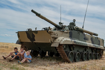 Three children in bright clothes are sitting in shade under modern battle tank. Children play until arrival of tankists. concept of contrasting innocent childhood and war