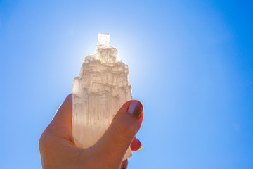 Person holding naturally carved mineral stone Selenite tower against sun and blue sky, stone has...