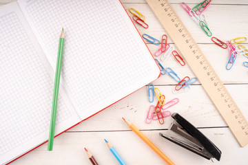 Various stationery placed on lumber table. From above colored pencils and open notebook arranged on wooden table near paper clips and stapler with ruler for studies