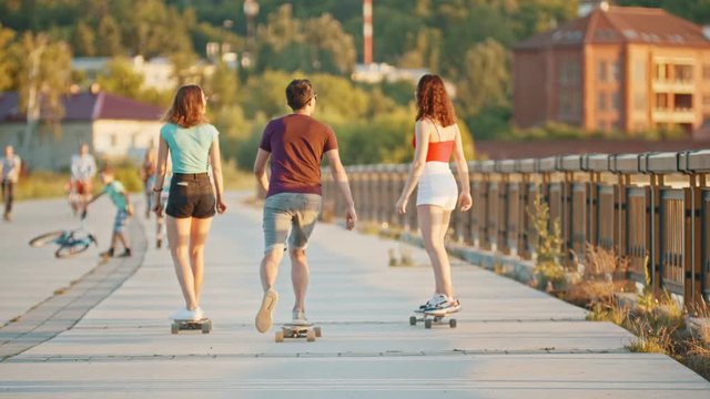 Two women and one man riding skateboards on a quay on a background of modern buildings on the sunset