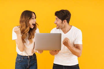 Portrait of successful couple man and woman in basic t-shirts rejoicing while standing together with silver laptop