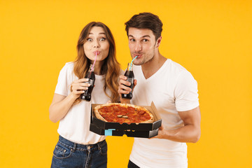 Portrait of joyful couple man and woman in basic t-shirts drinking soda beverage and holding pizza...