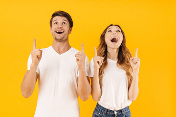 Portrait of content surprised couple man and woman in basic t-shirts pointing fingers upward at...