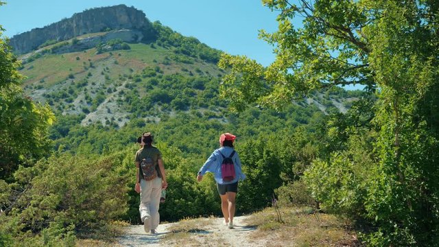 Two girls traveler girlfriend walk along the path and climb up the mountain plateau. They are surrounded by grass, shrubs and blue sky.