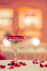 Greeteng card template. Red rose bud lies in a glass of champagne. Rose petals on the table. Bokeh background