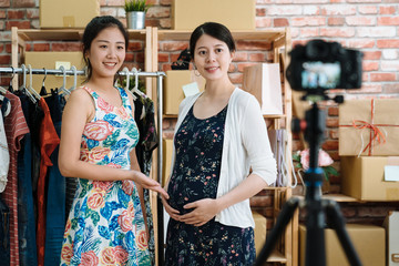 two beautiful girls showing selling on internet live stream online clothes shop. pretty woman with hand gesture showing introducing maternity dress comfort on pregnant woman model smiling to camera.