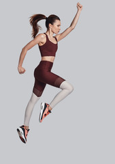 Fototapeta na wymiar Keep moving! Full length of attractive young woman in sports clothing jumping while exercising against grey background
