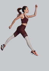 Flying high. Full length of attractive young woman in sports clothing jumping while exercising against grey background