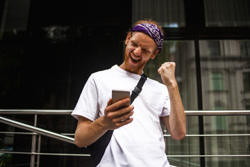 Naklejka premium Outdoor portrait of modern young man with mobile phone in the street. Excited man checking news on smart phone walking in the street. Win, winner concept. Hawing good news, wow surprised shocked face