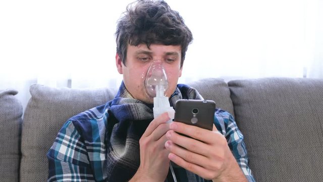 Use nebulizer and inhaler for the treatment. Sick man inhaling through inhaler mask and looking at mobile phone sitting on the sofa