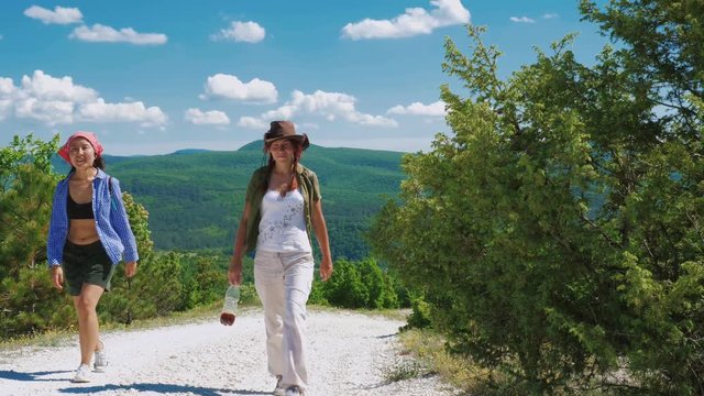 Two girls traveler girlfriend walk along the path and climb up the mountain plateau. They are surrounded by grass, shrubs and blue sky.
