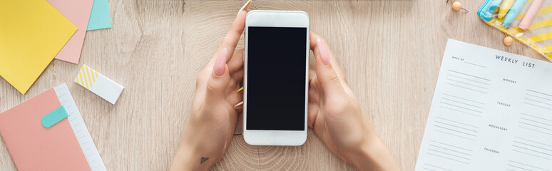 cropped view of woman holding smartphone over wooden table with laptop, weekly list and stationery