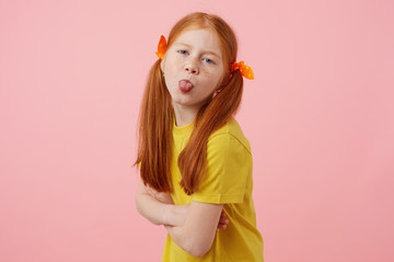 Portrait of petite freckles red-haired girl with two tails, looks and shows tounge at the camera,...