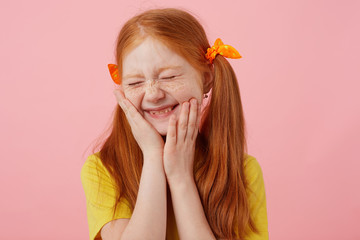 Portrait of smiling petite freckles red-haired girl with two tails, touches cheeks with closed eyes, wears in yellow t-shirt, stands over pink background. © timtimphoto