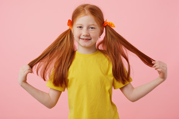 Happy petite freckles red-haired girl takes his two tails, broadly smiling and looks cute, wears in yellow t-shirt, stands over pink background.