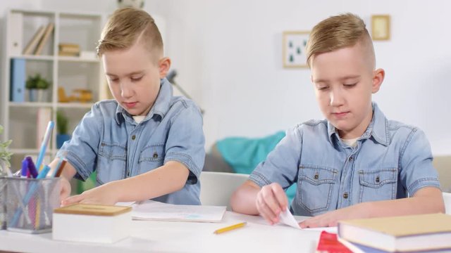 Chest-up shot of Caucasian identical twins with stylish haircuts, wearing denim shirts, sitting together at table and laboriously folding paper airplanes, throwing glances at each other&rsquo;s work