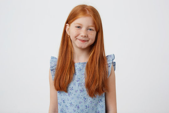 Portrait of petite cute calm smiling freckles red-haired girl wears in blue dress, stands over white background.