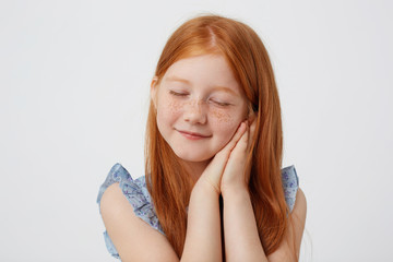 Portrait of dreaming little freckles red-haired girl in blue dress, looks sleppy, with folded palms at the cheeks, stands over white background.