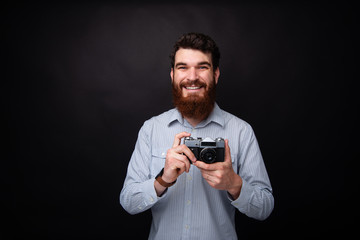 Let's make some photos. Young bearded man holding a vintage photo camera on black background.