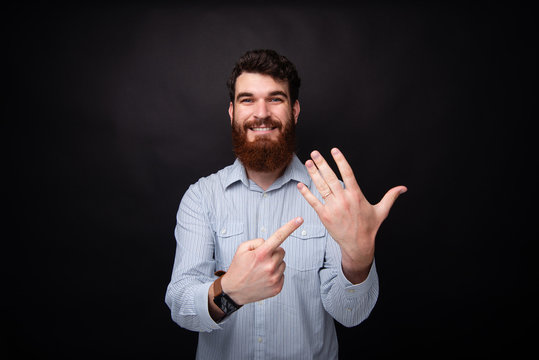 I&#039;m married! Young bearded man pointing at his marriage ring on his hand. Studio photo on black background.