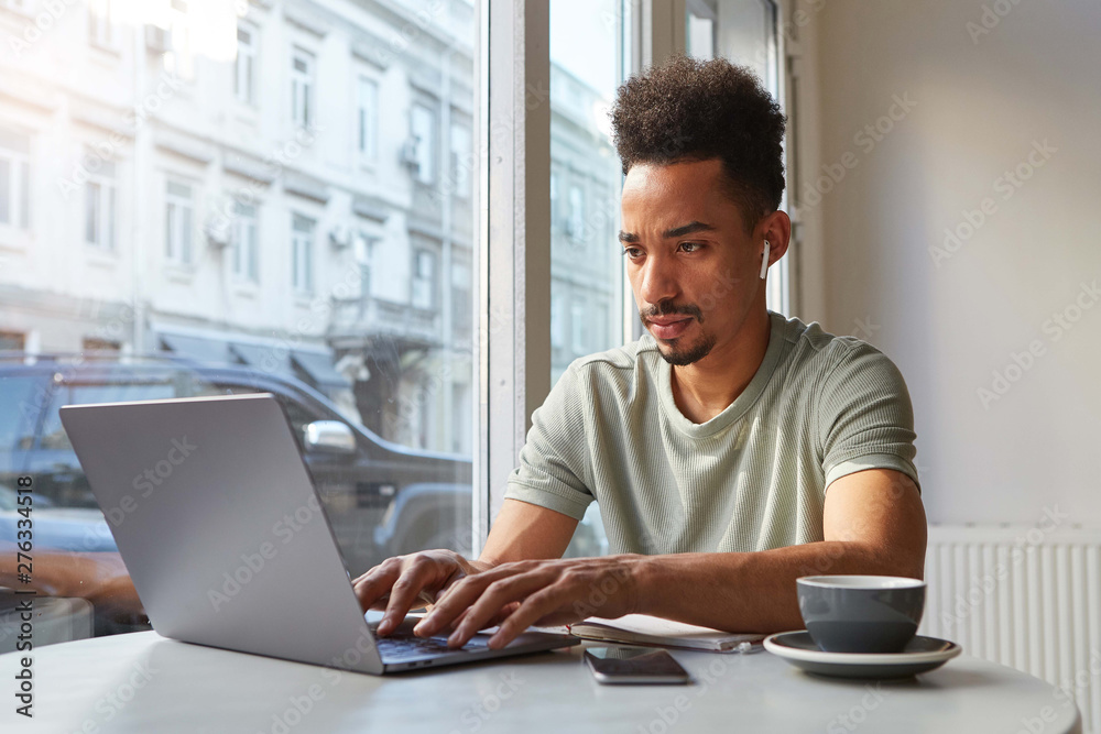 Wall mural portrait of concentrate young attractive african american boy, works at a laptop in a cafe, drinks c