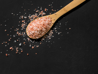 Fototapeta na wymiar Healthy spices concept. Large dark pink himalayan salt in a wooden spoon and spilled on a black table. Closeup
