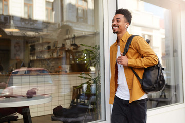 Photo of young happy African American man in yellow shirt walking down the street listening favorite song on headphones, looks cheerful, enjoy the sunny day in the city and broadly smiling. © timtimphoto