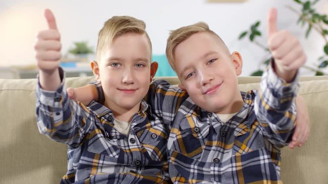 Waist-up portrait shot of 10-year-old Caucasian twin boys sitting together on sofa, hugging, looking straight, lifting up and extending their arms with thumbs up towards camera
