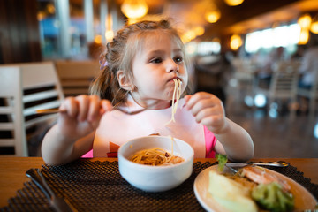 Litle girl is eating pasta in the restaurant.