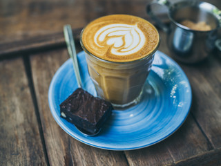 Piccolo Latte coffee topping with flower art from milk in small glass with a piece of homemade brownies cake on blue ceramic plate on wooden table