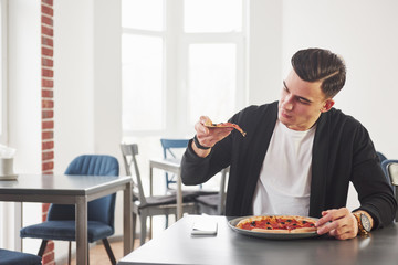 Looking at the slice. Having a breakfest with pizza at daytime. Young stylish man in black jacket
