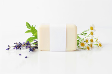 Obraz na płótnie Canvas Closeup of hand made herbal soap bar in blank paper label package. Mint leaves, lavender and feverfew flowers on white table backround. Spa concept. Skin product mockup scene. Cosmetic product.