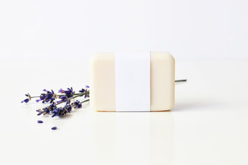 Closeup of hand made herbal soap bar in blank paper label package and bunch of lavender flowers on white table backround. Spa concept. Skin product mockup scene. Cosmetic product.