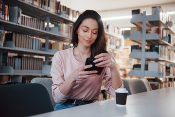 Using smartphone. Brunette girl in casual clothes having good time in the library full of books