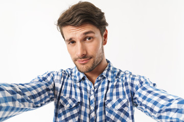 Serious young unshaved man dressed in casual shirt posing isolated over white wall background take a selfie by camera.