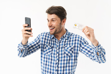Surprised handsome young man standing isolated over white wall background dressed in casual shirt using mobile phone holding credit card.