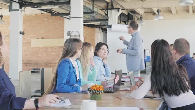 The male team leader conducts a presentation using a flipchart and communicates with colleagues. Employees sit at the table. Office life. Coworking. Modern interior in loft style