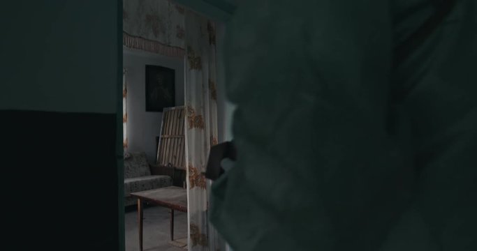 Man in a vintage WWII gas mask and Hazmat suit entering a cottage carefully looking around behind the door as he walks in and the dosimeter checks the level of radiation pollution. RAW video record.