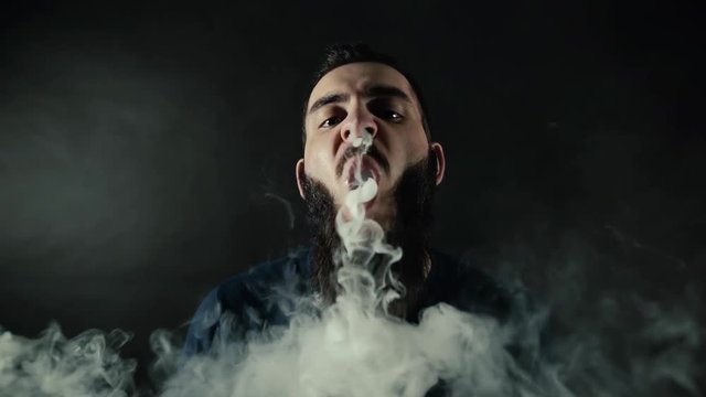 Close-up portrait of bearded guy exhaling big vapor ring from vape and  looks at the camera on black background in slow motion in professional dark  studio. Majestic art fog